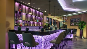 A bar area with a modern design, featuring a central bar counter, multiple seating options, and a variety of bottles and glasses on shelves. - Leonardo Royal Hotel Nürnberg (Nuremberg)
