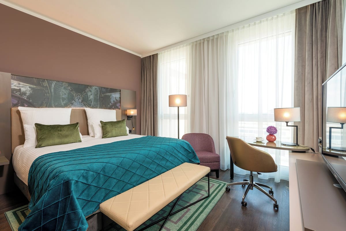 Quarto de hotel com uma cama grande, uma escrivaninha com cadeira, uma luminária e vista para a cidade pela janela.