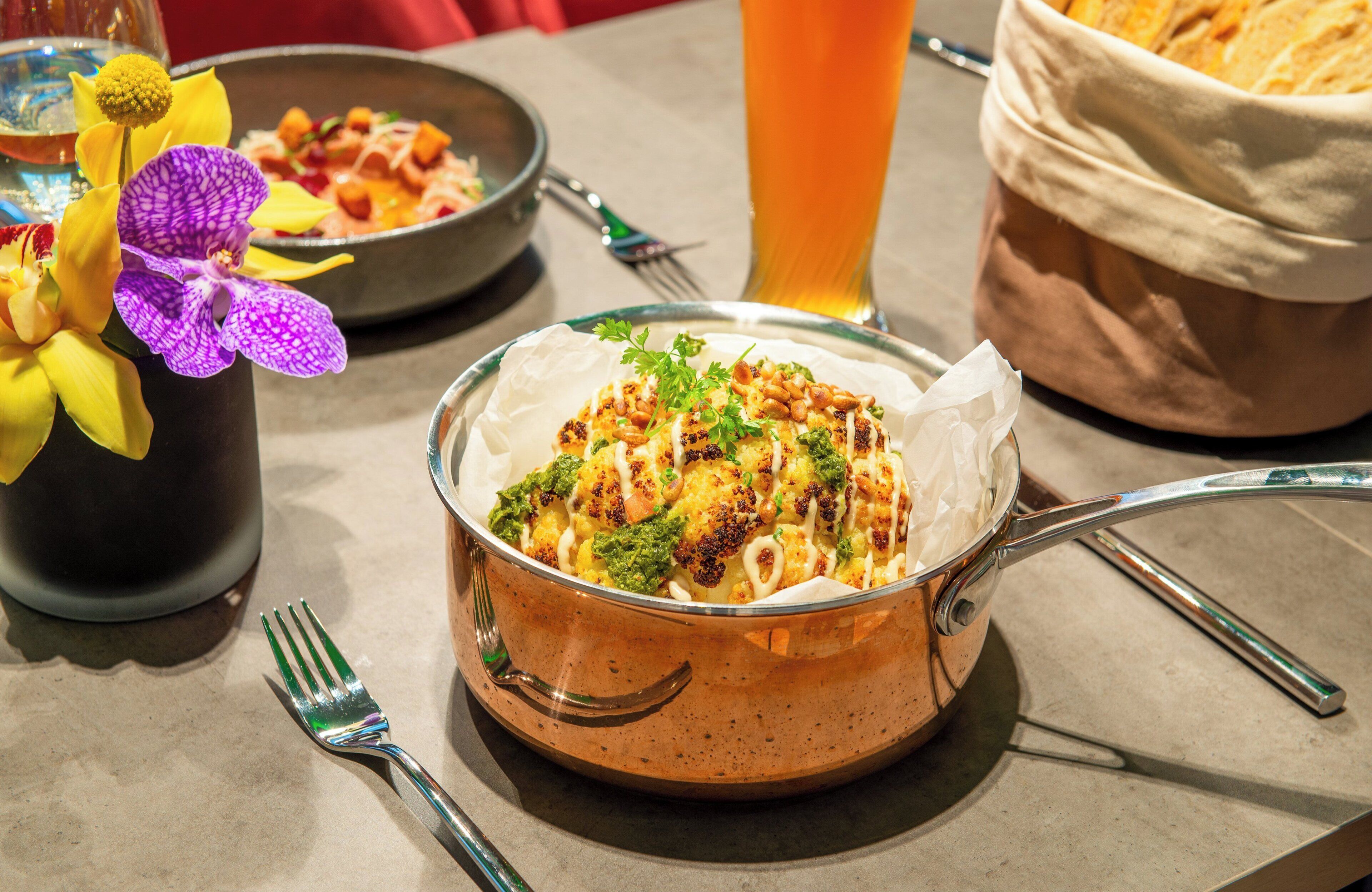 A dining table with a meal in a copper pot, a glass of beer, and a basket of bread.