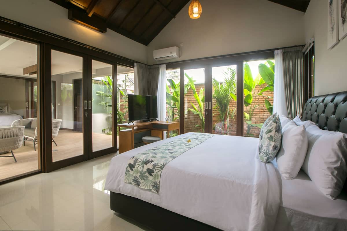 A bedroom with a bed, a desk, a television, and a sliding glass door leading to a balcony with plants.