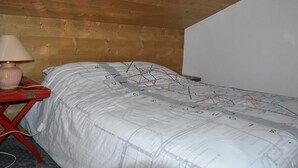 Une chambre avec un plafond en bois, un lit recouvert d’une couette à motifs de cartes géographiques, une table rouge avec une lampe et une prise de courant.