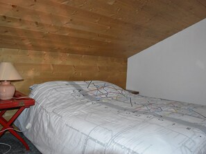 Un dormitorio con techo de madera, una cama con edredón con diseño de mapa, una mesa roja con una lámpara y un enchufe.
