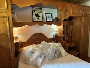 Une chambre à coucher avec une tête de lit en bois, un lit recouvert d’une couverture moelleuse, une table de chevet avec une lampe et une étagère sur laquelle se trouvent un tableau encadré et un petit vase.