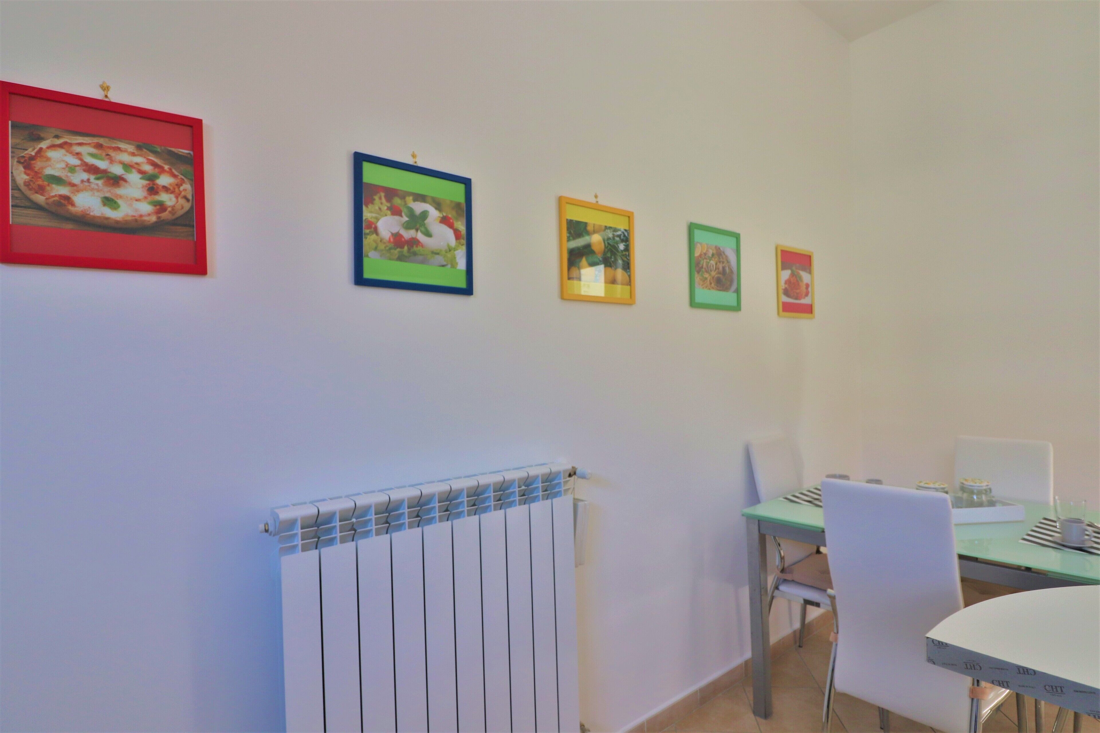 A dining area with a table, chairs, and a radiator. There are four framed pictures on the wall.