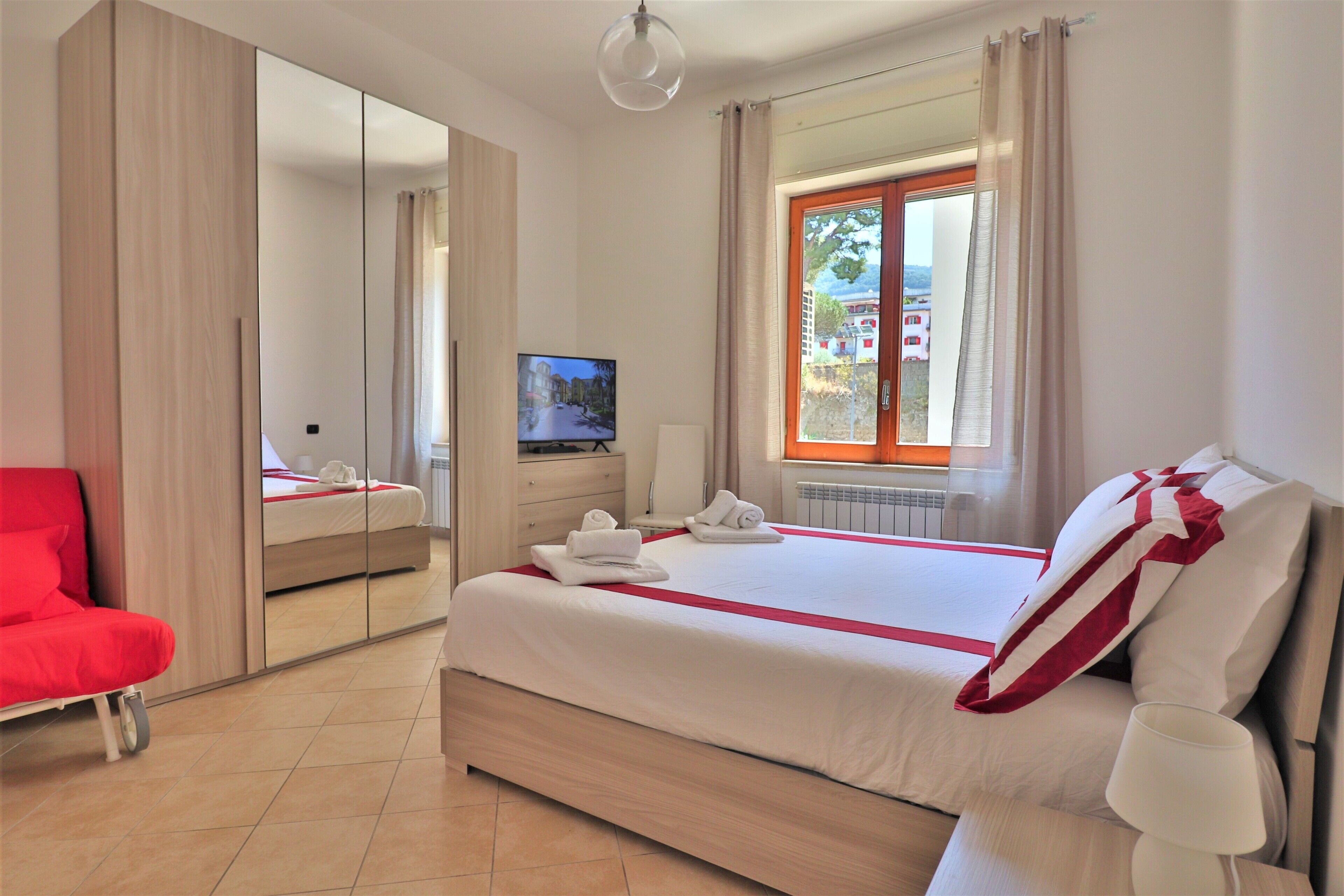 A hotel room with a large bed, a television, a red chair, and a window with curtains.