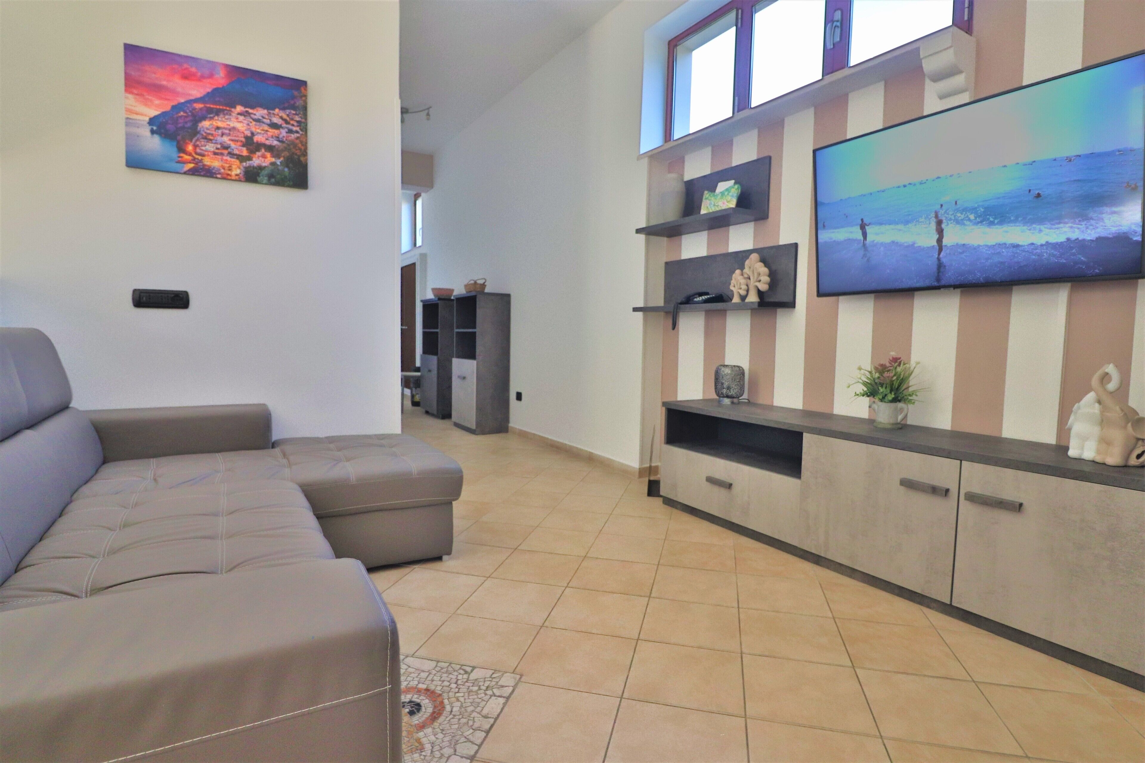Een woonkamer met een hoekbank, een flatscreen televisie bevestigd aan een gestreepte muur en een klein kastje met decoratieve objecten.