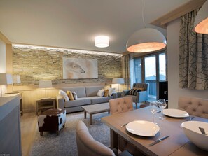 Ett modernt vardagsrum med en vägg av sten, en soffa, ett matbord för två personer och ett stort fönster med gardiner.