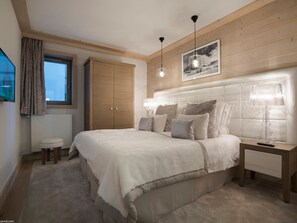 A bedroom with a large bed, a wooden wardrobe, a window with curtains, a bedside table with a lamp, and a framed picture on the wall.