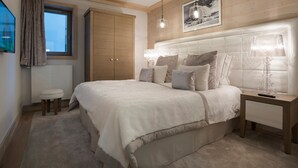 Ein Schlafzimmer mit einem großen Bett, einem Holzschrank, einem Fenster mit Vorhängen, einem Nachttisch mit Lampe und einem gerahmten Bild an der Wand.