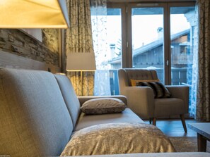 A living room with a sofa, a chair, a lamp, and a view through a glass door to the outside. - Modern apartment in Courchevel 1550 with electric box and wifi (Courchevel 1550)