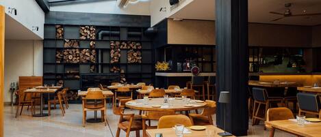A spacious restaurant with round wooden tables and chairs, a high ceiling with skylights, and a black wall with shelves of wood logs.
