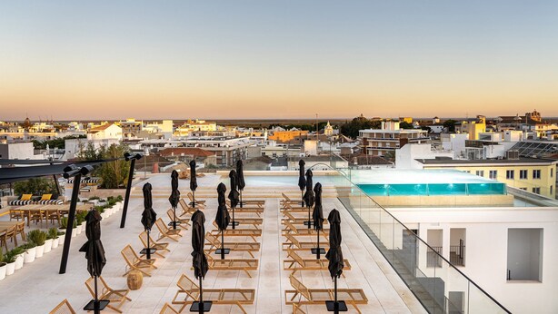 A rooftop terrace with sun loungers and umbrellas, a swimming pool, and a cityscape view.
