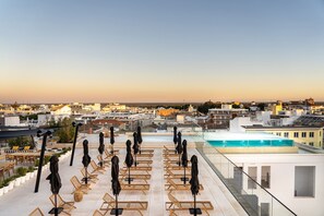 A rooftop terrace with sun loungers and umbrellas, a swimming pool, and a cityscape view.