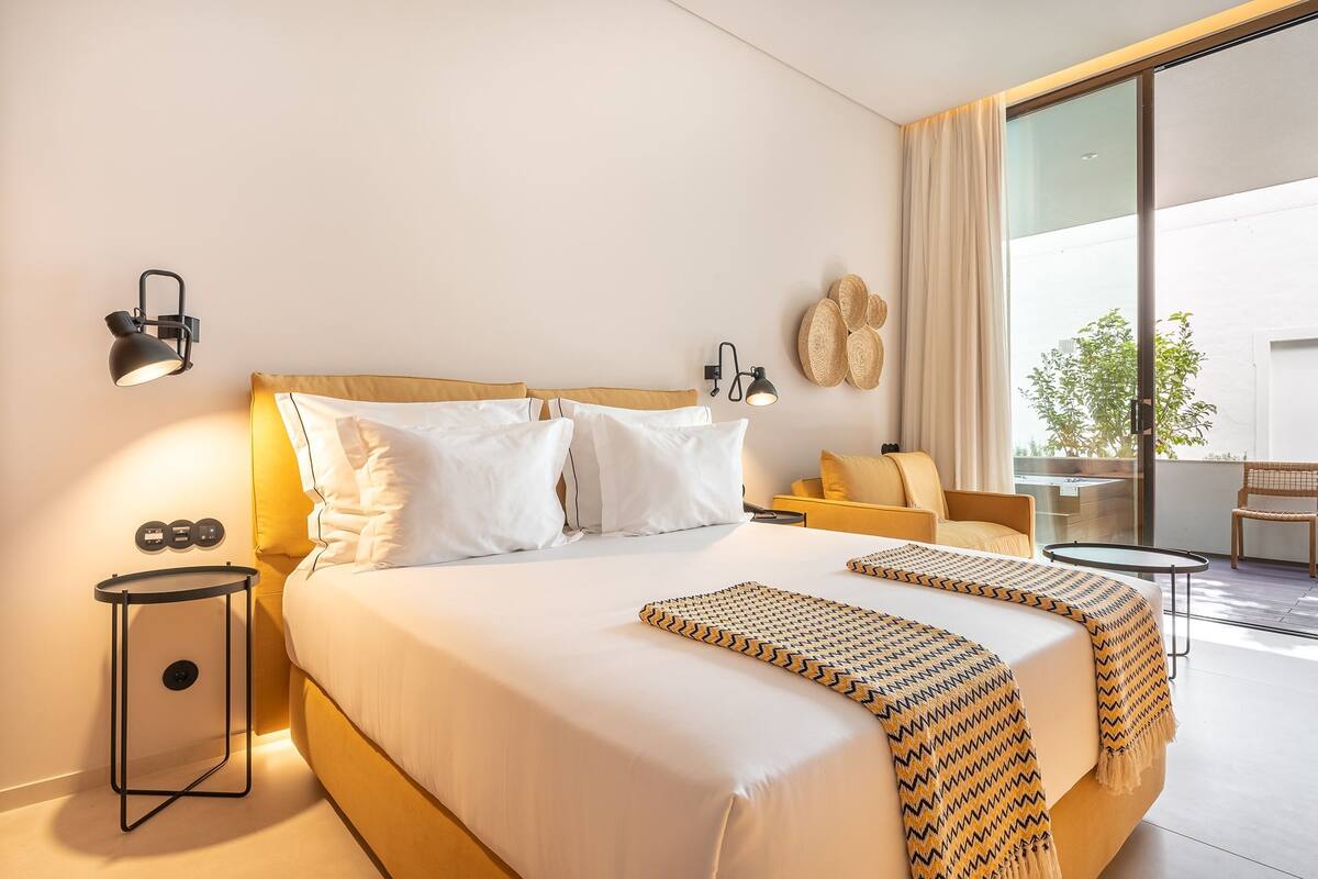 A hotel room with a large bed, two bedside lamps, a small side table, a sofa, and a balcony with a view.