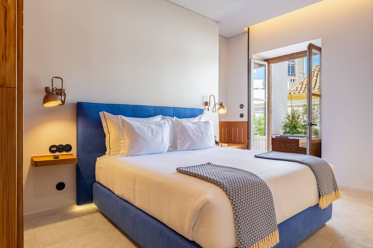 A neatly made bed with a blue headboard, white bedding, and a patterned throw blanket. There are two bedside tables with lamps, a small shelf with decorative items, and a view of the outside balcony.