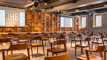 A conference room with rows of wooden chairs and tables, wooden shelving units, and large screens displaying "3HB HOTELS".