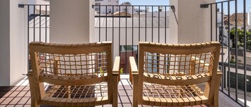 Two wooden chairs with a woven backrest on a balcony with a metal railing, overlooking a cityscape with buildings and a clear sky.