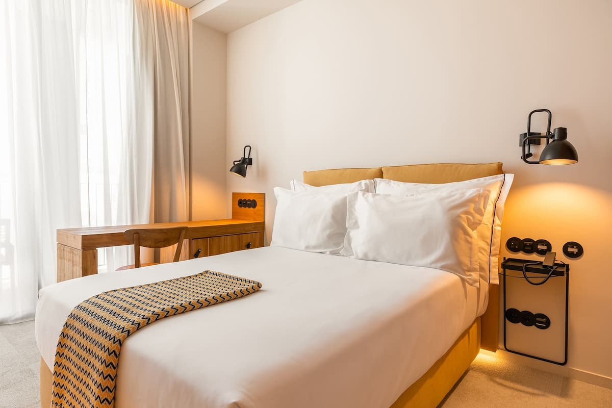 A hotel room with a bed, bedside table, lamp, and a wall-mounted shoe rack.