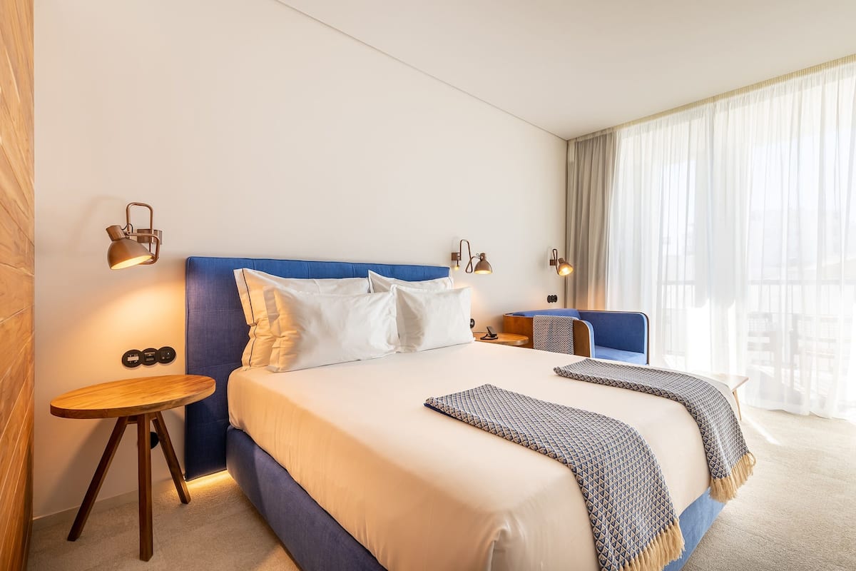 A hotel room with a large bed, two bedside tables, a small desk, and a large window with sheer curtains.