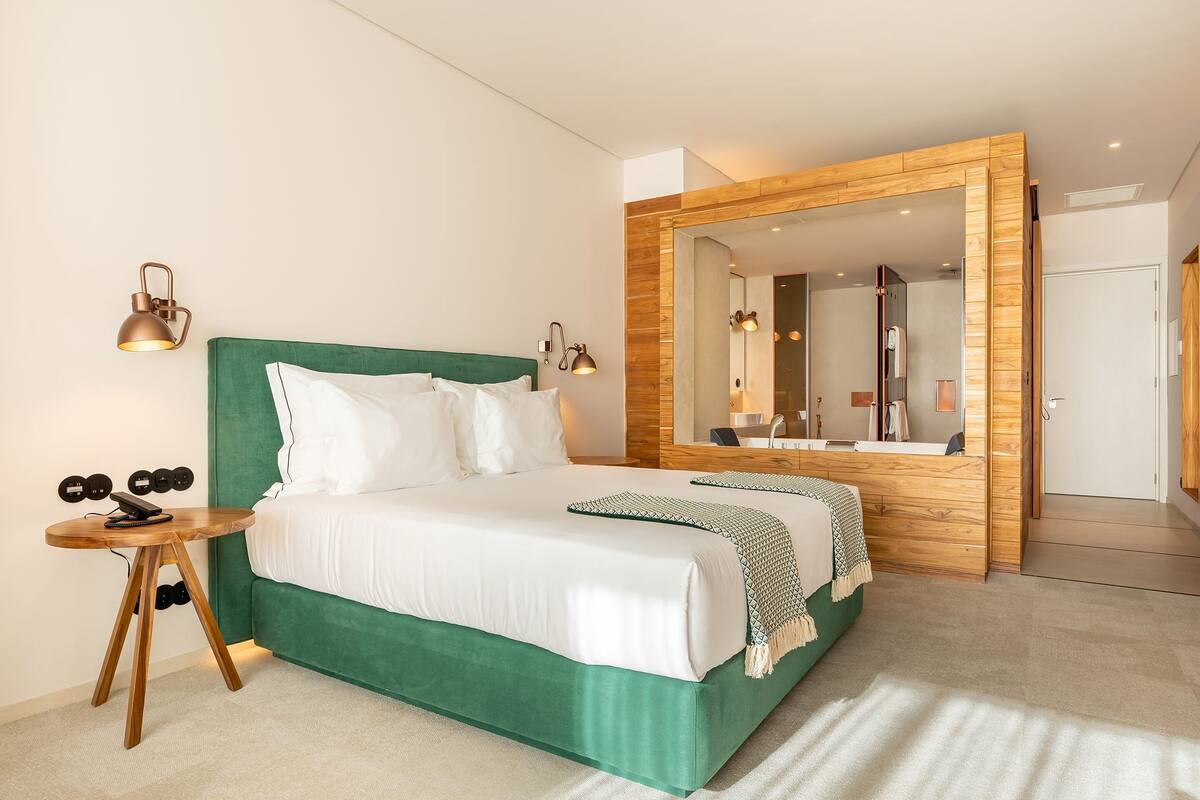 A modern hotel room with a green upholstered bed, a wooden side table with a lamp, and a large mirror with a wooden frame.