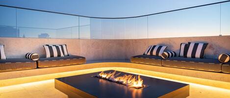 A modern outdoor seating area with a built-in fire feature, cushioned seating with striped pillows, and a glass balustrade.
