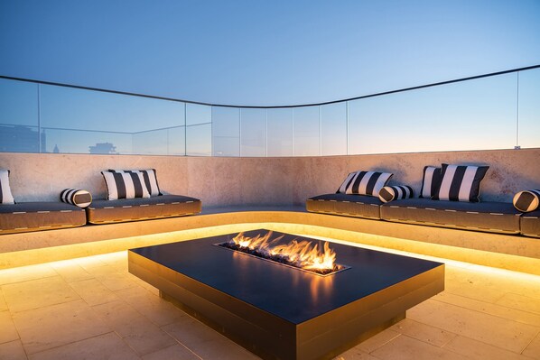 A modern outdoor seating area with a built-in fire feature, cushioned seating with striped pillows, and a glass balustrade.