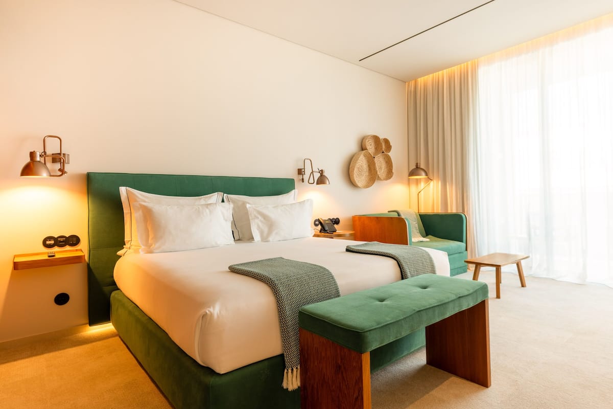 A modern hotel room with a large bed, a green upholstered bench, a small wooden table, and a wall-mounted lamp.
