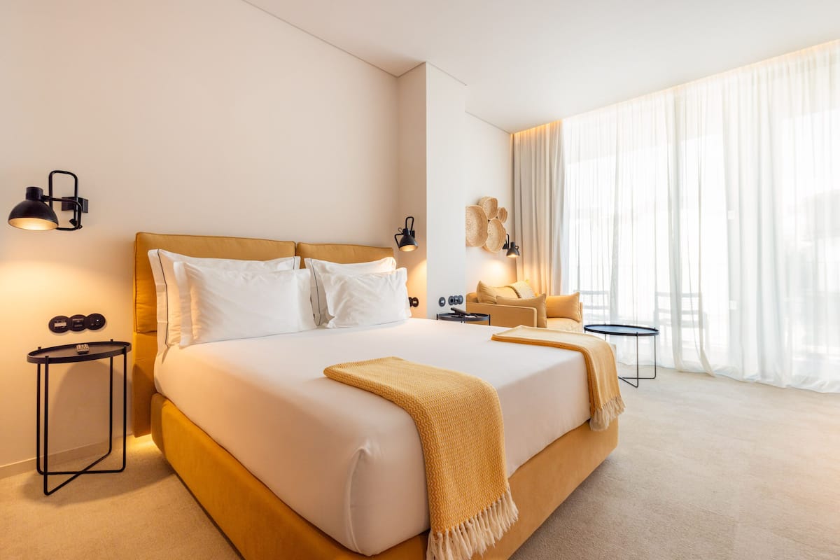 A hotel room with a large bed, bedside tables, a small seating area, and a large window with sheer curtains.
