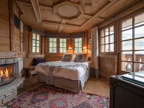 A cozy room with a fireplace, a bed with pillows, a patterned rug, and large windows with curtains.