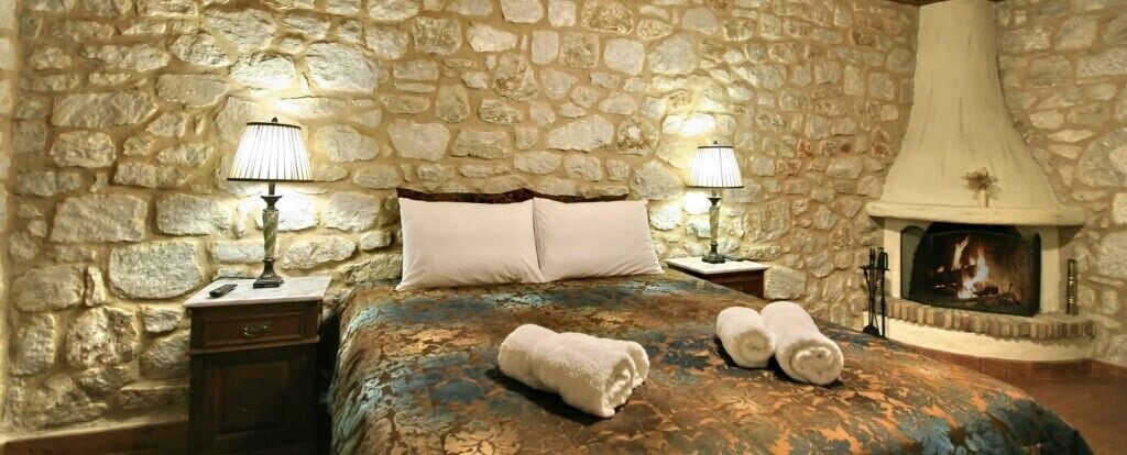 Una habitación con pared de piedra, una cama con colcha estampada, dos lámparas de noche, una chimenea y toallas enrolladas sobre la cama.