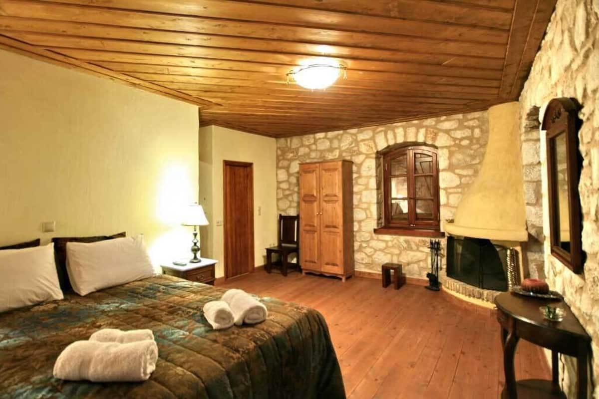 Une chambre confortable avec un plafond en bois, des murs en pierre, un lit recouvert d’une courtepointe, une table de chevet, une lampe, une cheminée en pierre et une armoire en bois.
