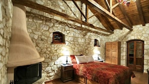 A stone-walled bedroom with a large fireplace, wooden beams, a bed with red bedding, and two bedside lamps. - Archontiko Kolokotroni (Gortynia)