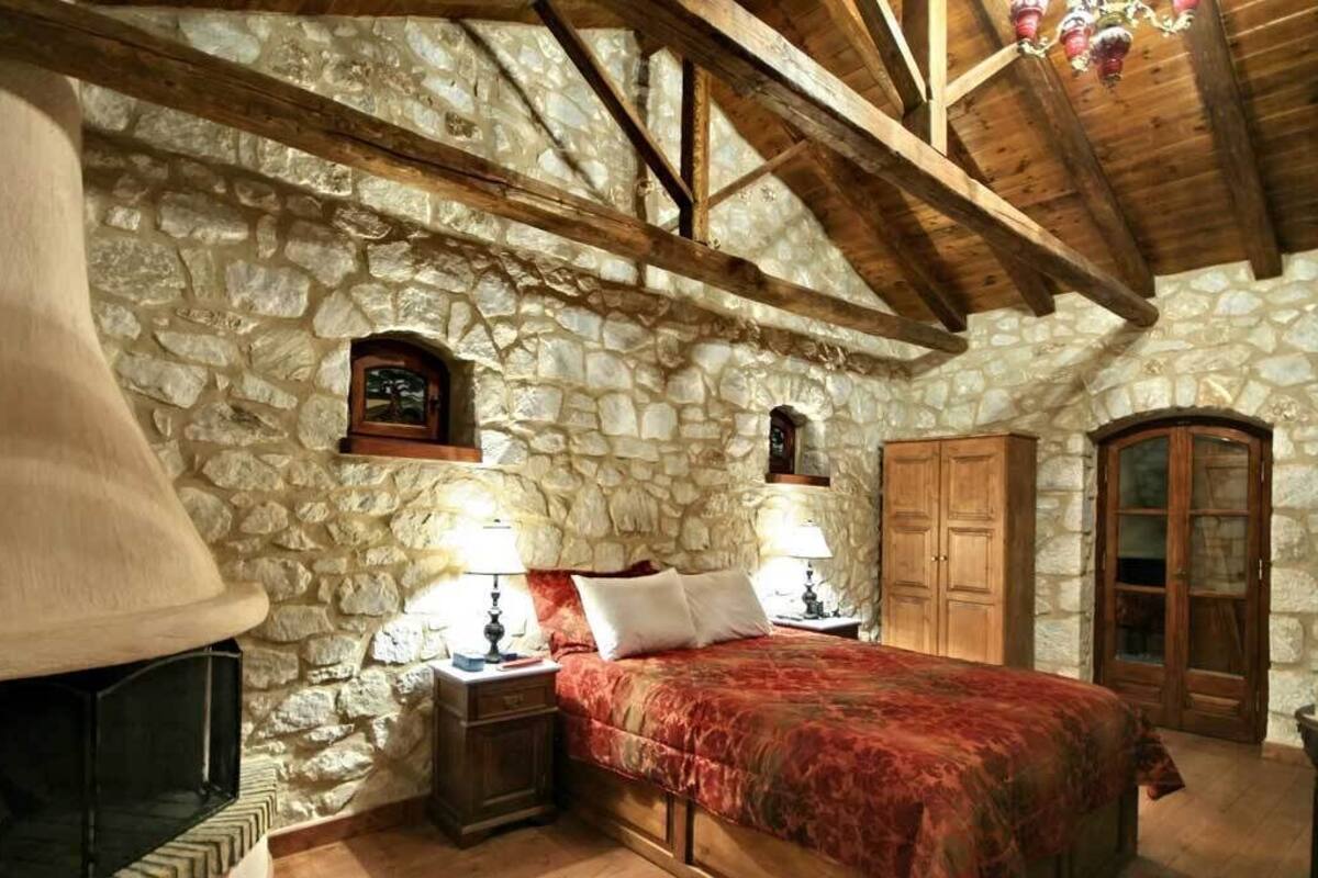 Une chambre aux murs de pierre, dotée d’une grande cheminée, de poutres apparentes, d’un lit avec une literie rouge et de deux lampes de chevet.