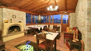 A cozy room with a stone fireplace, wooden ceiling, and large windows offering a view of the mountains. - Archontiko Kolokotroni (Gortynia)