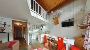Ein geräumiger Wohnbereich mit hoher Decke, einem Esstisch mit roten Stühlen, einer Küche im Hintergrund und einer Treppe, die in ein Obergeschoss führt.