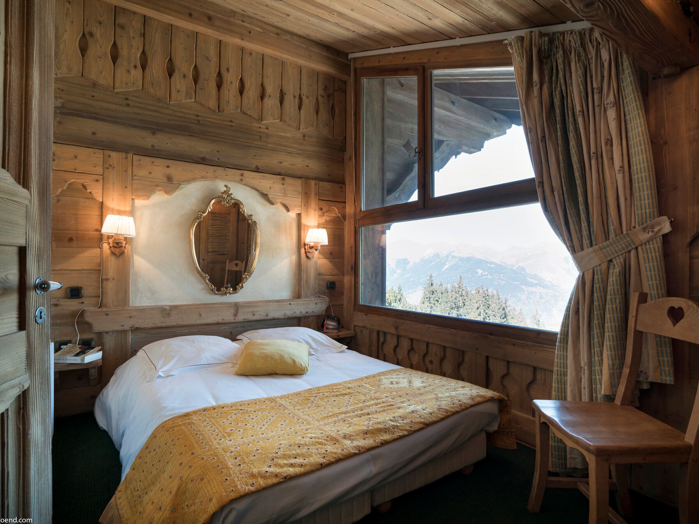 A wooden cabin bedroom with a bed, a chair, a mirror, and a window with a mountain view.