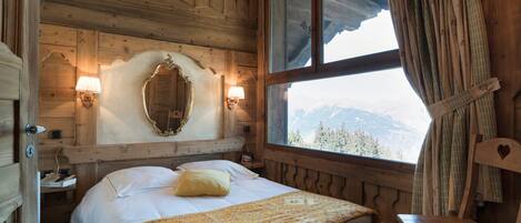 A wooden cabin bedroom with a bed, a chair, a mirror, and a window with a mountain view.