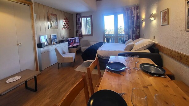 Ett mysigt rum med en säng, ett matbord för två personer, en tv och ett fönster med utsikt över träd.