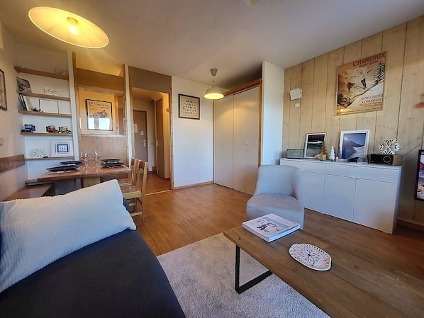 Un salon moderne comprenant un canapé, une table basse et un coin repas avec une table et des chaises. On y trouve également un meuble blanc sur lequel sont posés une radio et des objets décoratifs.