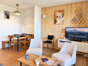Un salon avec un mur en bois, un coin repas avec une table et des chaises, un téléviseur diffusant un paysage de montagne et une affiche encadrée.