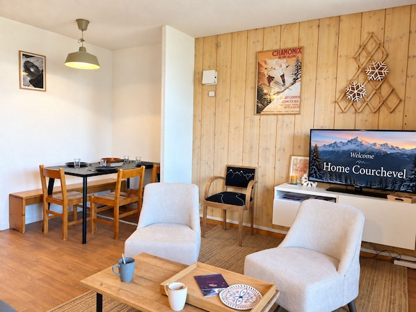 A living room with a wooden wall, a dining area with a table and chairs, a television displaying a mountain scene, and a framed poster.