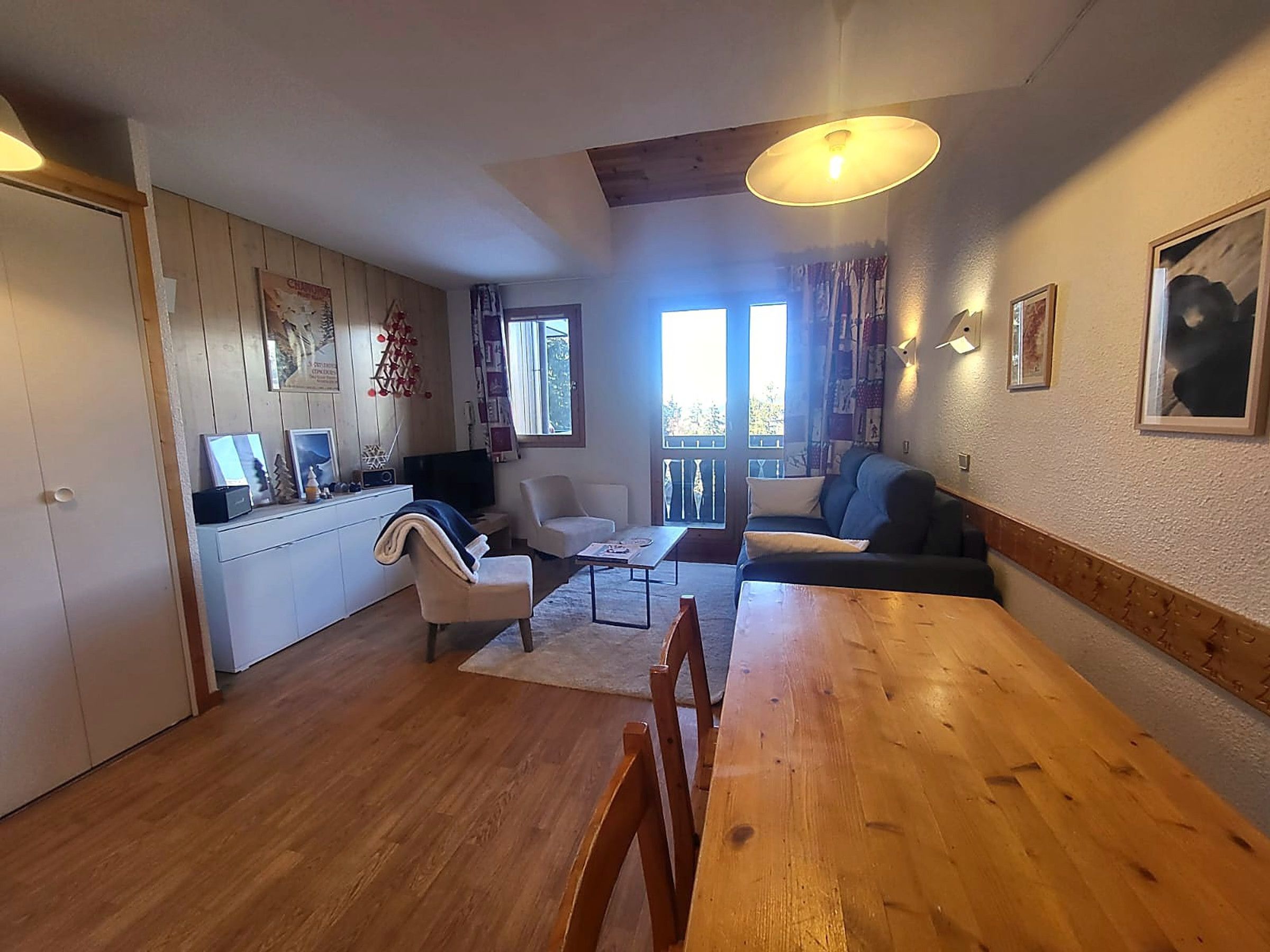 A cozy living room with a wooden dining table, a sofa, and a TV. There is a kitchen area with cabinets and a window.