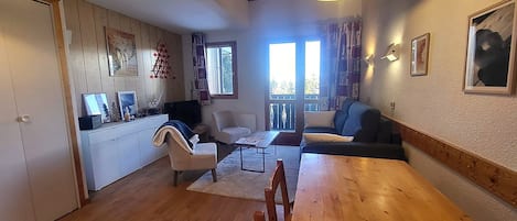 A cozy living room with a wooden dining table, a sofa, and a TV. There is a kitchen area with cabinets and a window.