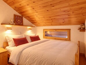 A bedroom with a wooden ceiling, a bed with white bedding and red pillows, a window, and a teddy bear on a shelf.