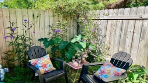 A wooden fence with climbing plants and flowers, two black chairs with floral cushions, and a small table with a vase and glasses.