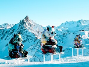 Trois grandes sculptures de pandas, assises sur des tables blanches, avec en arrière-plan une montagne enneigée.