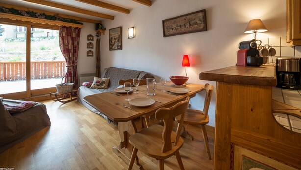 Un comedor acogedor con una mesa de madera preparada para cuatro personas, un sofá y una zona de cocina con cafetera.