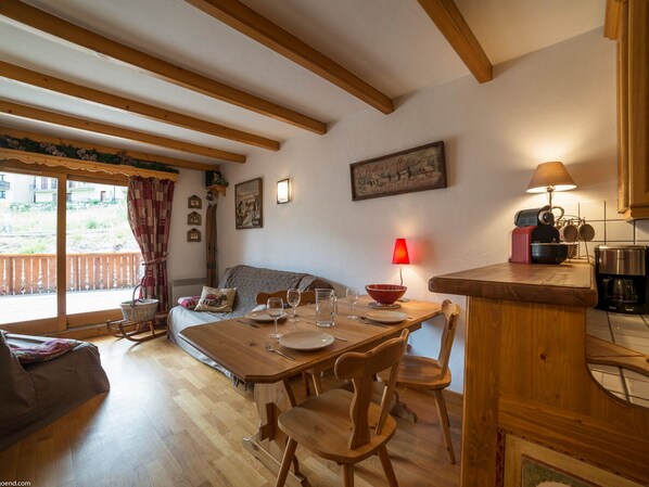 Un coin repas chaleureux avec une table en bois dressée pour quatre personnes, un canapé et un espace cuisine équipé d’une machine à café.