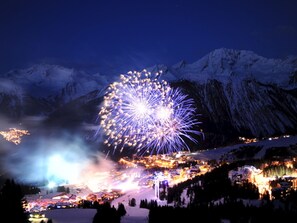 Un paysage montagneux enneigé, avec une ville en contrebas et un grand feu d’artifice illuminant le ciel.