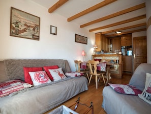 Un salon chaleureux avec un canapé, des fauteuils, une table à manger, des poutres en bois, un coin cuisine et un tableau encadré au mur.